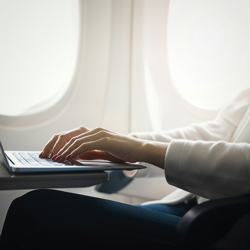 Person typing on a laptop resting on a tray table by airplane window, hands poised over keys, light streaming through rounded window shapes.