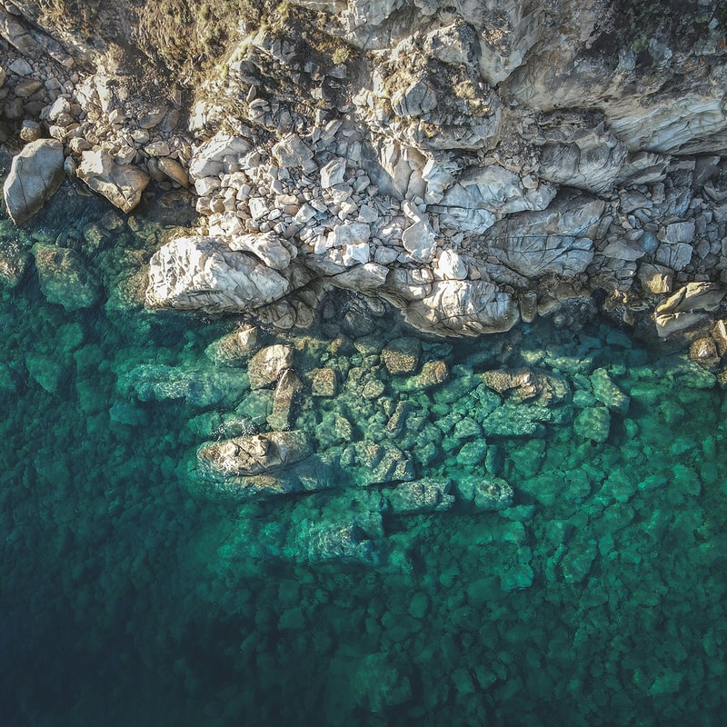 Aerial view of a rocky coastline with gray jagged stones transitioning into clear, teal-green water; rocks extend into the sea, forming a rugged shoreline.