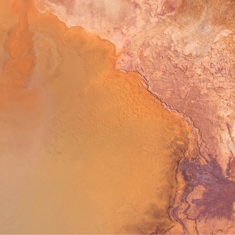 Aerial view of a desert shoreline with orange-brown sand and cracked, drying crust forming from a pale to deep orange gradient along a coastal edge.