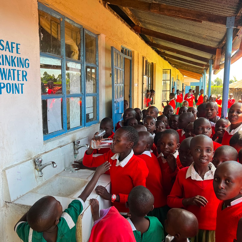 Crowded school water point with many students in red uniforms queueing at blue-framed sinks under a covered walkway outside a beige building.