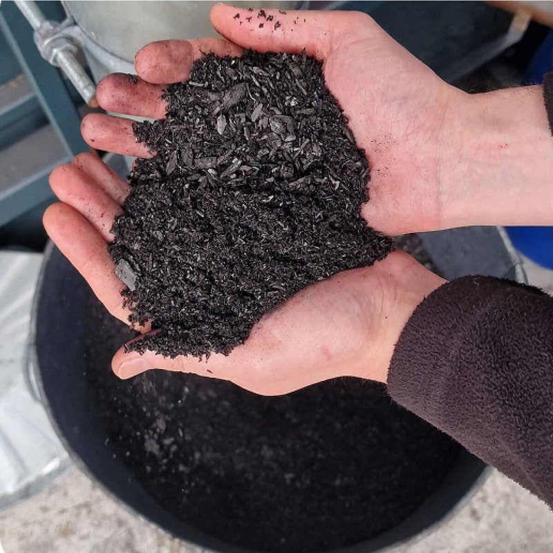 Dark crushed black soil or compost held in two hands over a container, dirty fingertips, industrial setting in background.