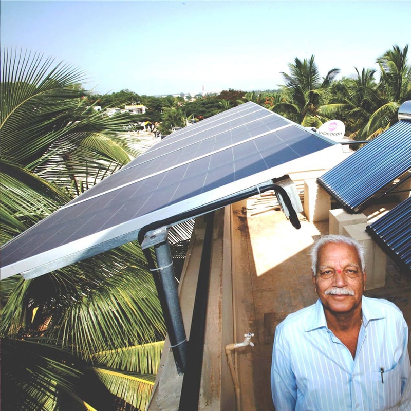 Solar panels on a rooftop arching over a man with white hair in a light blue striped shirt, tropical palm trees, and a bright blue sky in the background.