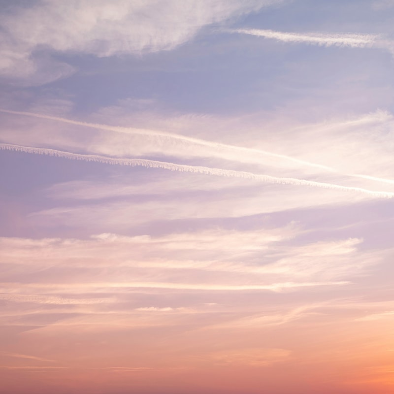 Vibrant sunset sky with soft pastel hues of pink, purple, and orange, wispy clouds and contrails slicing diagonally across the calm horizon.