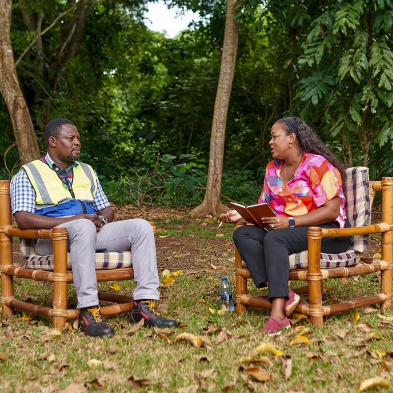 Two adults sit on wooden chairs outdoors in a green, leafy park; a man in a safety vest and a woman in a colorful blouse discuss, with a notebook and bottle nearby.