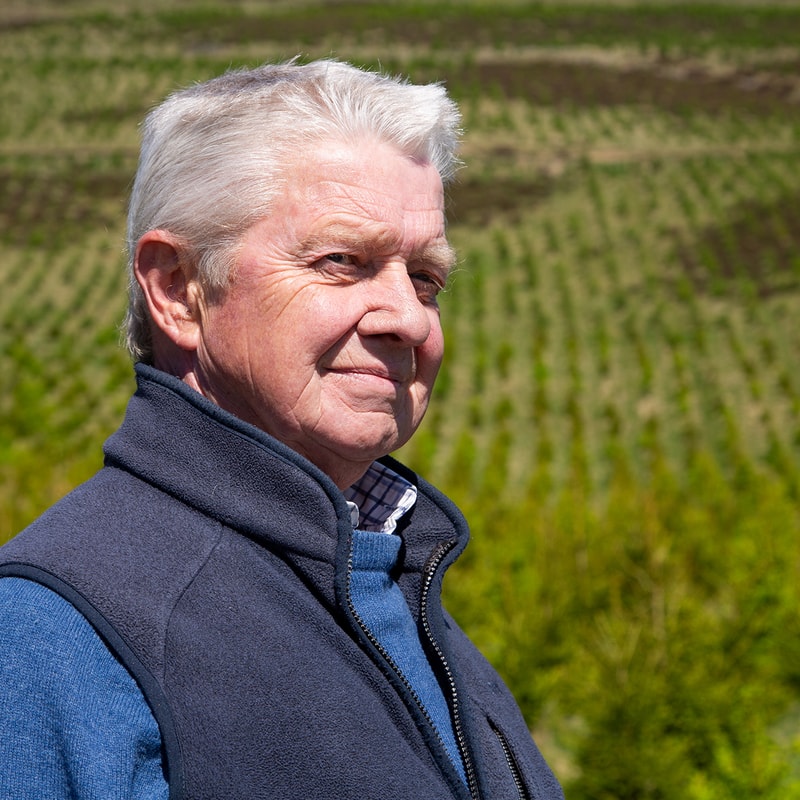 An older person with short white hair smiling outdoors, wearing a navy fleece vest over a blue sweater, with a green field in the background.