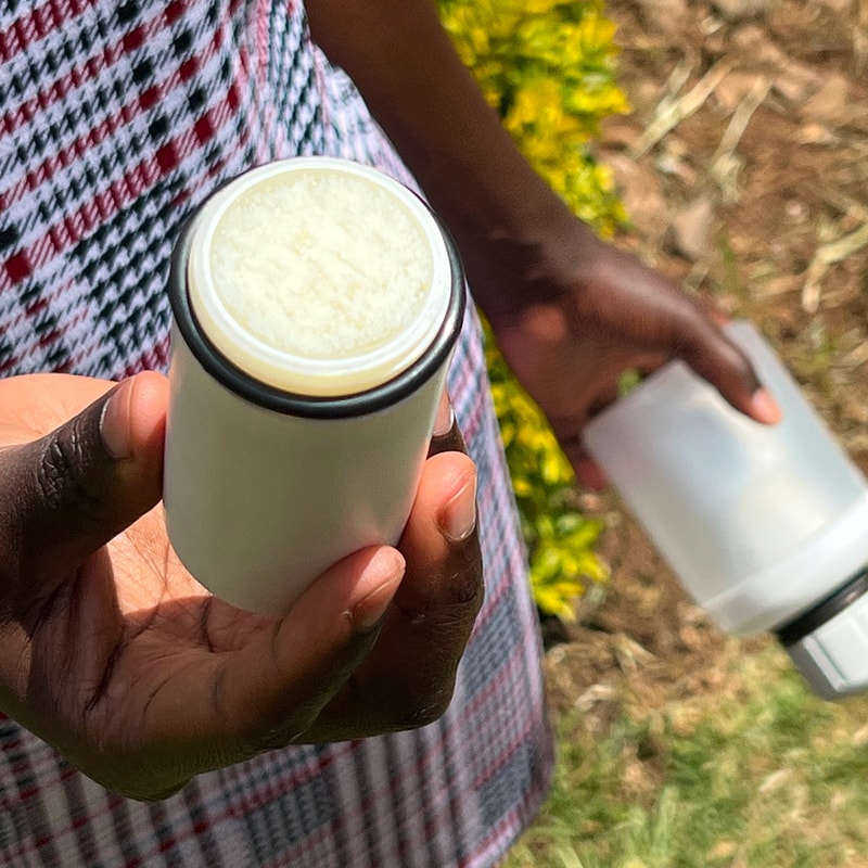 Person outdoors holding a white insulated cup with a frothy light-colored beverage, lid off, wearing a patterned garment; background shows greenery and soil.
