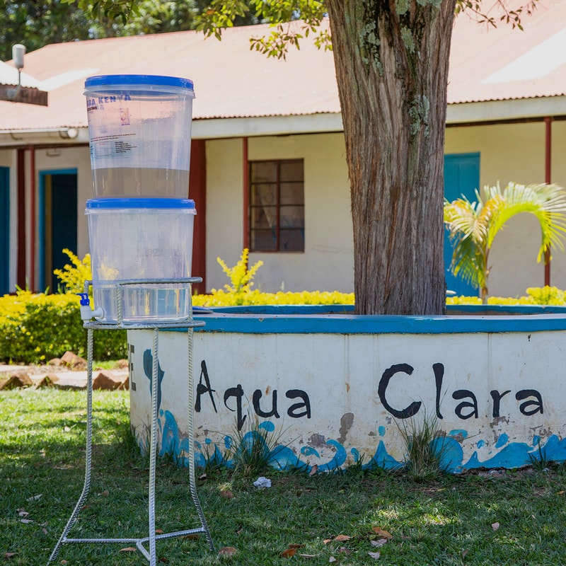 Two stacked transparent water containers with blue lids on a metal stand beside a painted circular planter containing a tree, in front of a building with yellow flowers and a red roof.