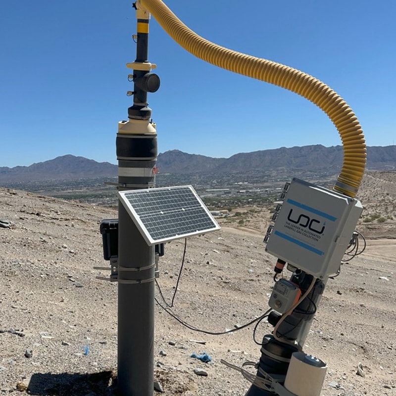 Field equipment on a rocky hillside with a tall vertical mast, solar panel, and yellow insulated hose, set against a desert valley and distant mountains.