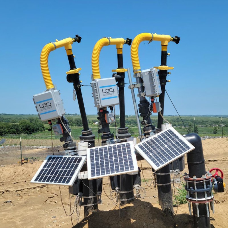 Multiple yellow and black industrial pipes with attached control boxes rise from the ground, with solar panels angled in front and a rural landscape in the background.