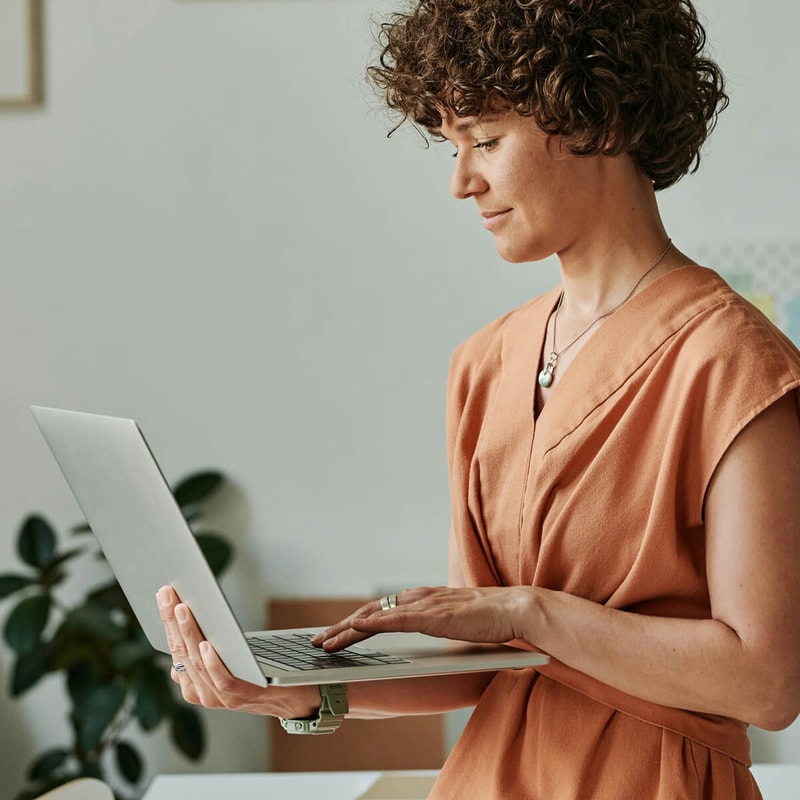 Person with short curly hair in a rust-colored top stands at a desk, holding an open silver laptop and typing with both hands, indoors.