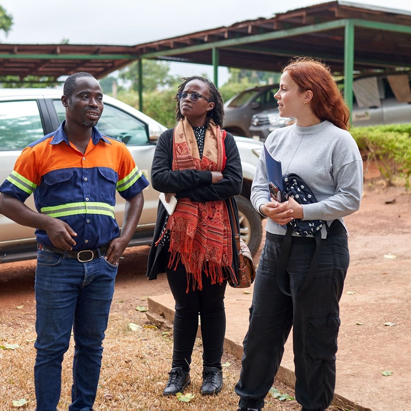 Three people stand outdoors near a vehicle: a man in an orange and blue work shirt speaking, a woman with glasses and a red scarf listening, and another woman with red hair holding a notebook and bag.