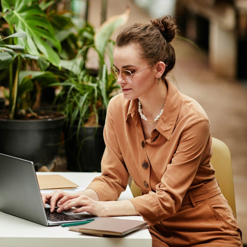 A person wearing a peach-colored button-up dress and pearl necklace sits at a white table, typing on a laptop outdoors with green potted plants in the background.
