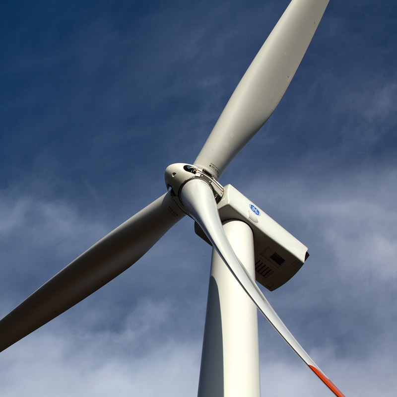Close-up of a wind turbine nacelle and rotor hub with blades extending against a blue sky, showing the connection and housing at the top of the tower.