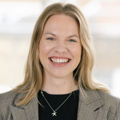 Smiling woman with shoulder-length blond hair wearing a plaid blazer and a black top, indoors with a bright, blurred background.
