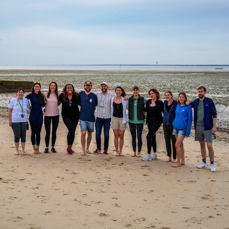 Group of about 12 people standing together on a sandy beach near low tide, posing for a photo with arms around each other, ocean and sky in the background.