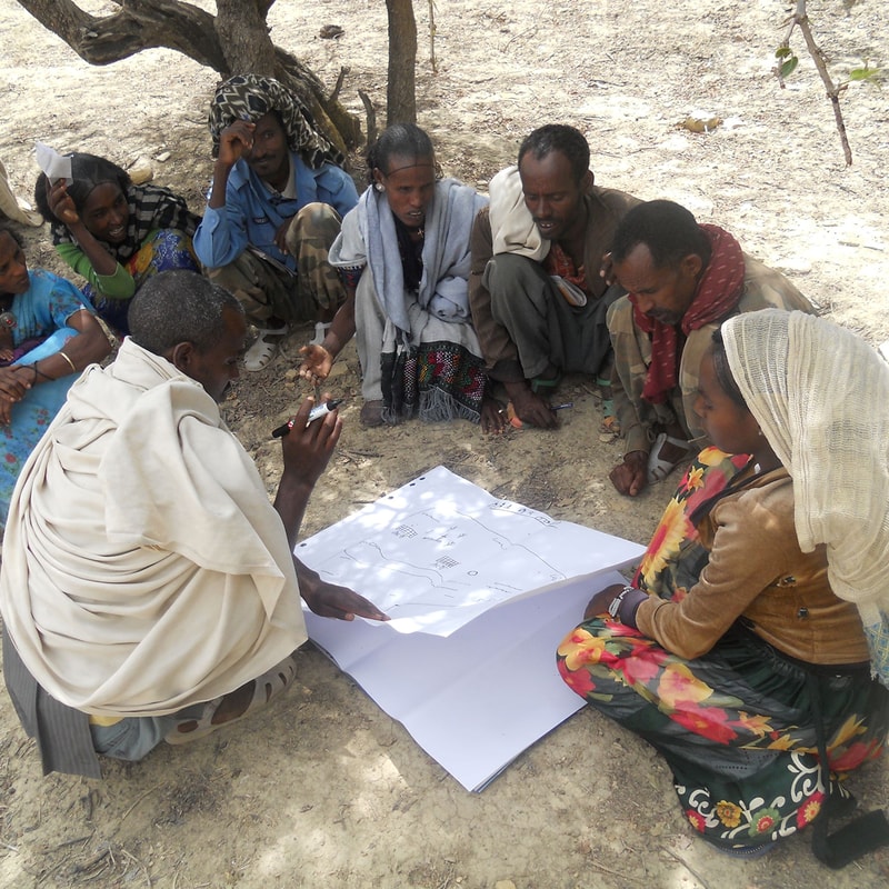 Group of people sits on dry ground under a tree, gathered around a large sheet of paper with drawings; a man in a white robe points and writes while others watch.