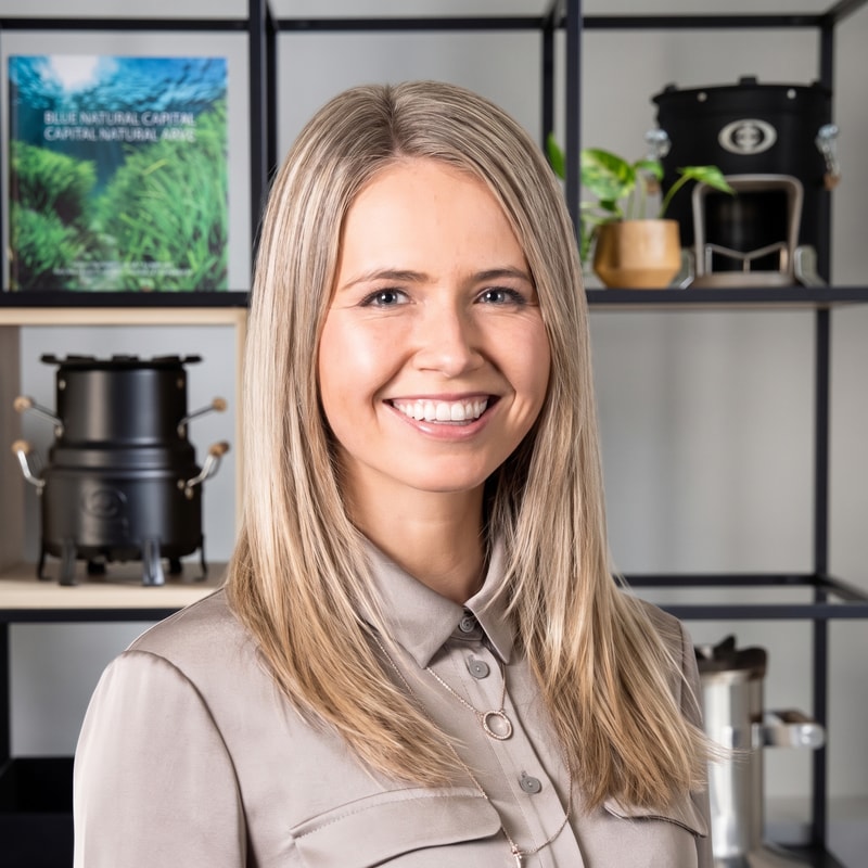 Smiling woman with long blond hair wearing a beige button-up shirt, standing in front of a shelf with coffee equipment and plants.