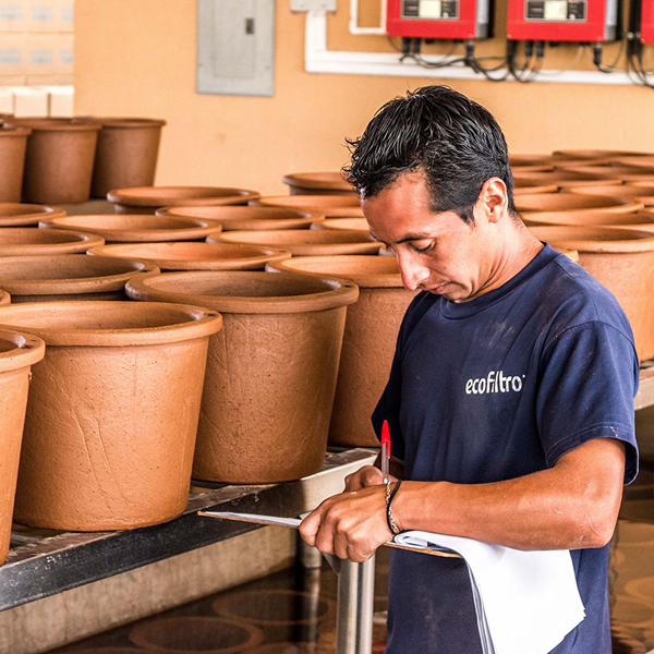 A man wearing a dark blue shirt with “ecoFTRo” logo inspects and writes on a clipboard in a room filled with large brown clay pots arranged on shelves.