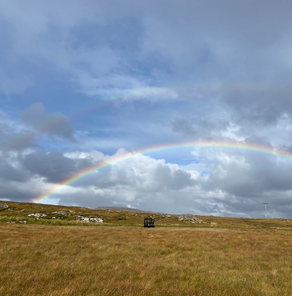 Vast grassy field with a lone dark vehicle near the horizon, under a dramatic cloudy sky and a bright rainbow arching across the landscape.