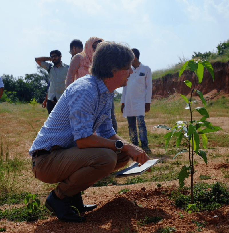 A man in a blue striped shirt crouches next to a young plant, holding a notebook, while a group of people stands in the background on a sunny outdoor field.