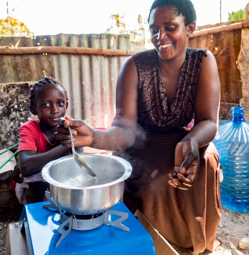 A smiling woman and a young girl cook over a small blue gas stove outdoors; the woman stirs a steaming pot while the girl watches, in a corrugated metal surroundings.