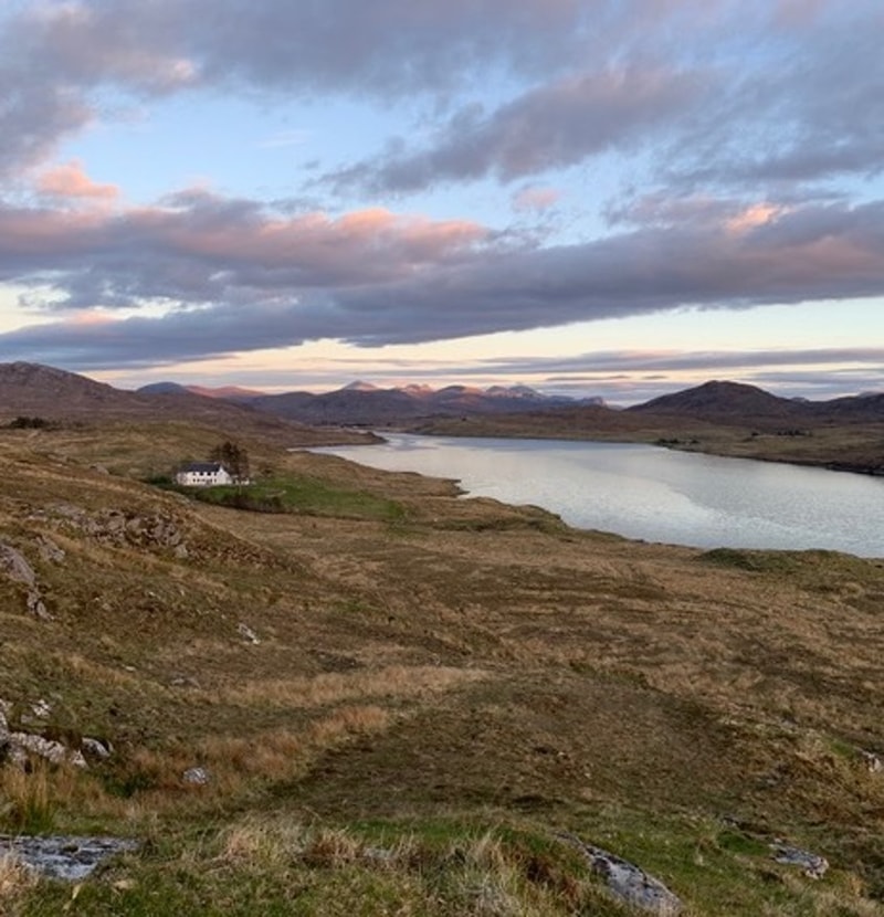 Scenic moorland with a winding river, a small white house by the shore, rolling hills, and a soft pink-tinged sunset sky above distant mountains.