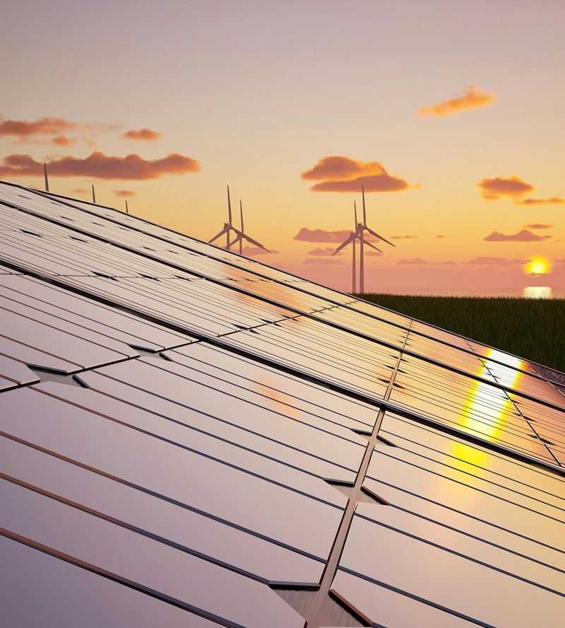 Rows of solar panels on a sloped field reflect a warm sunset, with distant wind turbines and a calm sea horizon under a pastel sky.