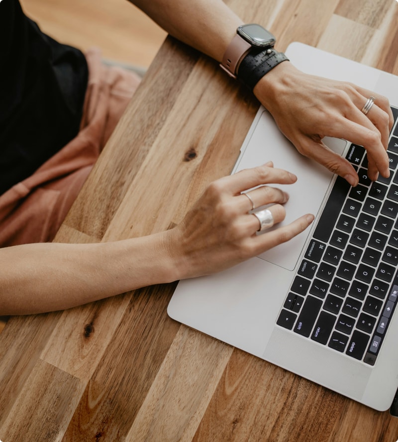 A person sits at a wooden table typing on a silver laptop, wearing a smartwatch and multiple rings, with their hands positioned over the keyboard and trackpad.