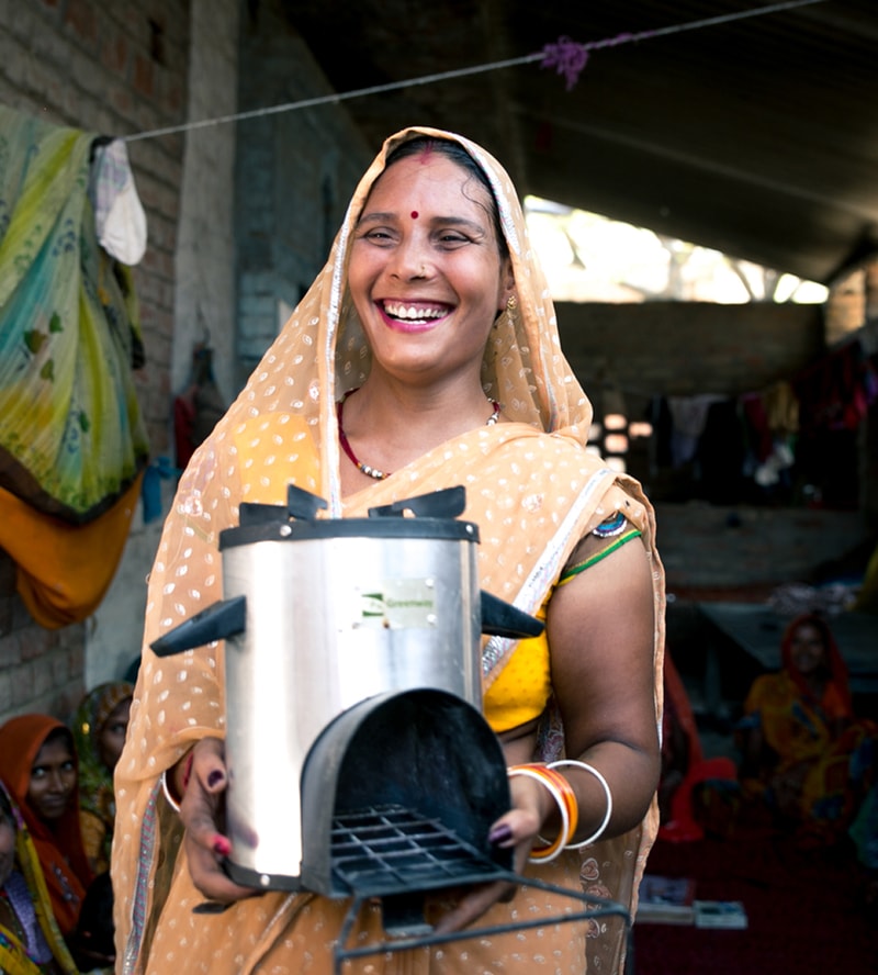Smiling woman in a peach saree holds a metallic kitchen appliance with a handle and drip tray, standing in a sunlit open-air room with clothes hanging in the background.