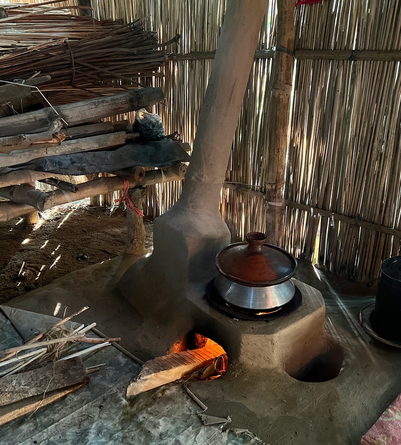A clay stove with a tall chimney inside a thatched-willow hut, a pot on top, fire glowing from an opening beneath, and stacked wood and tools along the wall.