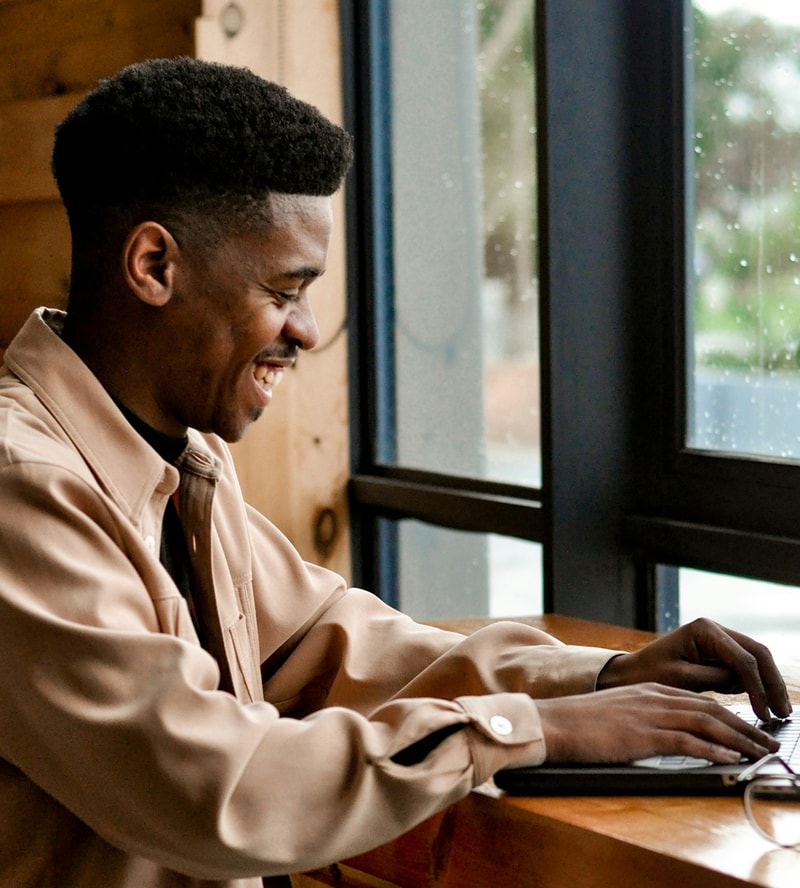 A person with short black hair sits at a wooden table by a window, smiling while typing on a laptop. Rainy glass and natural light visible outside.