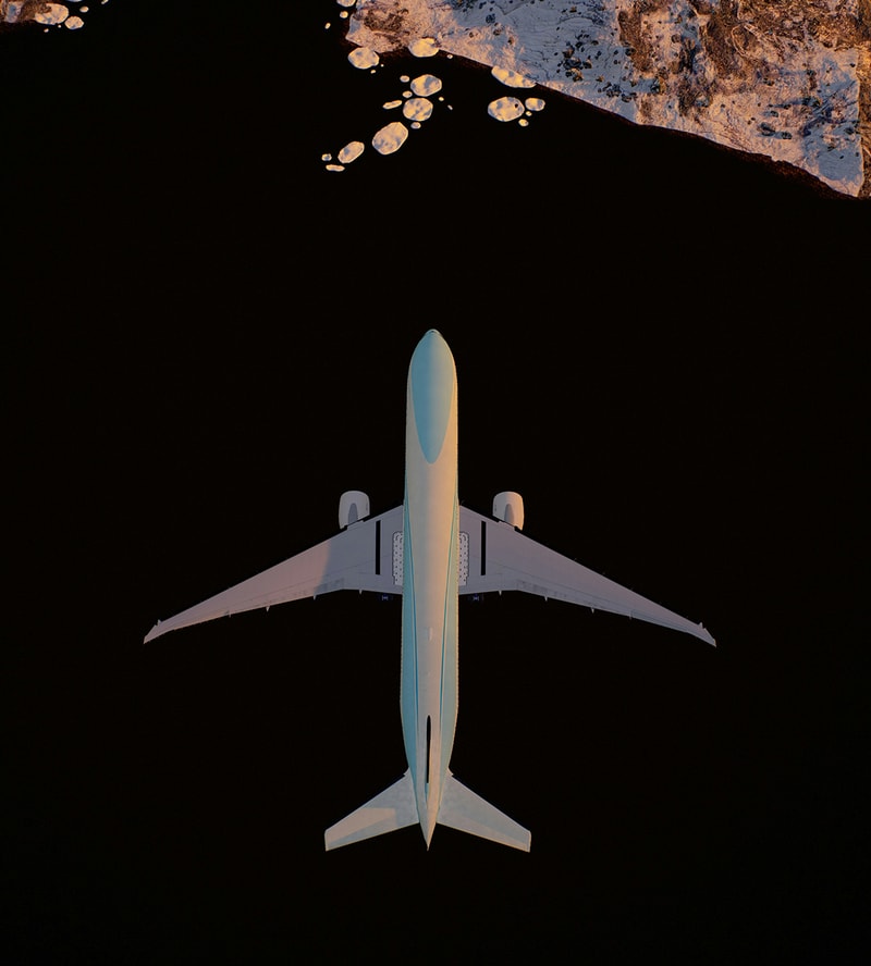 Heavy white airliner viewed from above against dark background, with wings spread and two engines, approached by rocky shoreline at top edge.