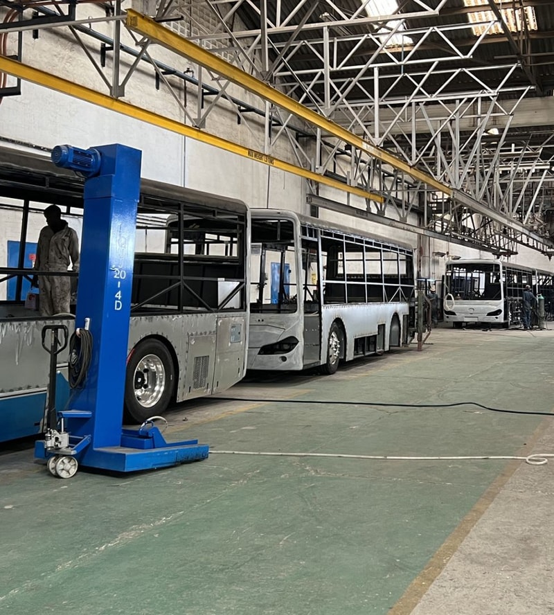 Inside a bus maintenance hangar, silver buses are lined up beside a blue vehicle lift, with workers inspecting and tools hanging on the wall.