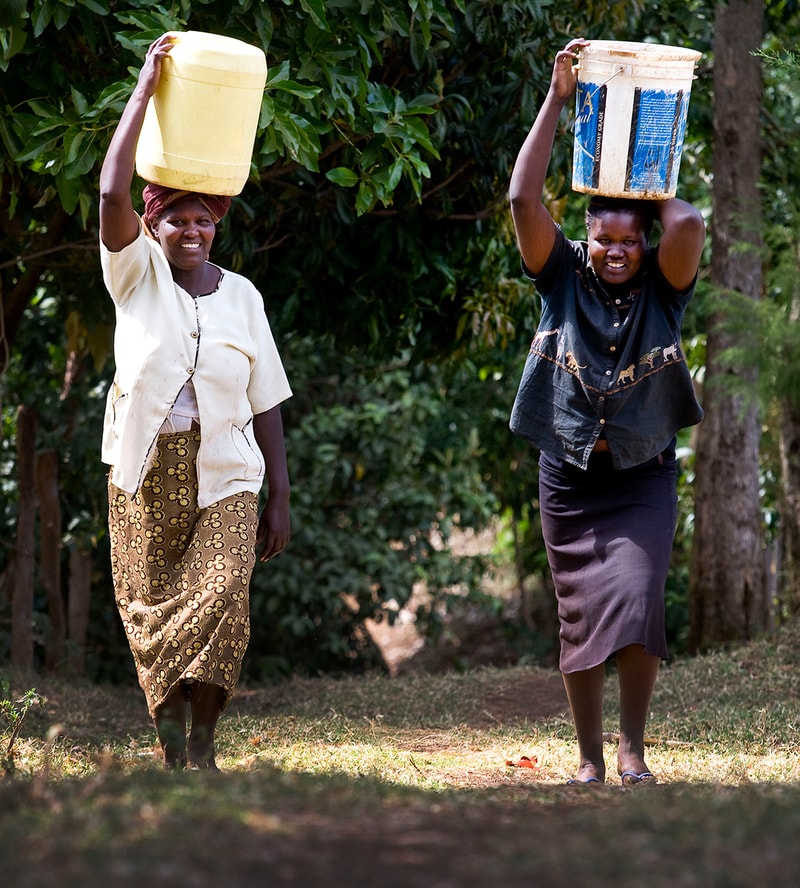 Two women walk along a leafy path carrying large containers on their heads, one in a light cardigan and patterned skirt, the other in a dark shirt and knee-length skirt.