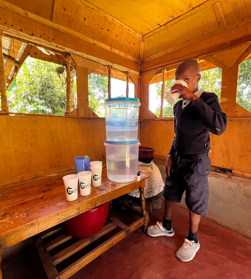 Child wearing dark clothing drinks from a cup beside a large water dispenser in a wooden shelter with windows and green foliage outside.