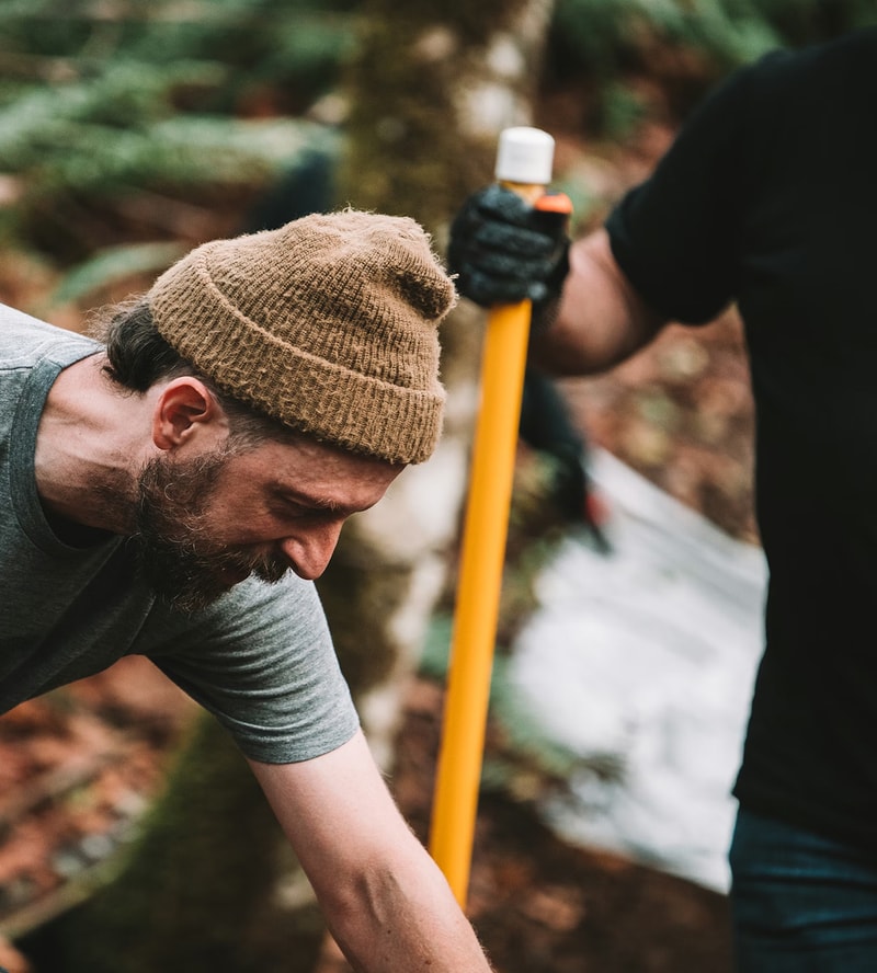 A bearded person wearing a tan knit cap and gray shirt bends forward outdoors, while another person in a black shirt holds a yellow tool handle in the background.