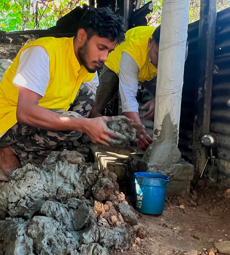 Two workers in yellow vests mix and apply cement around a vertical pipe, building a small concrete structure beside a blue bucket in an outdoor, earthy setting.
