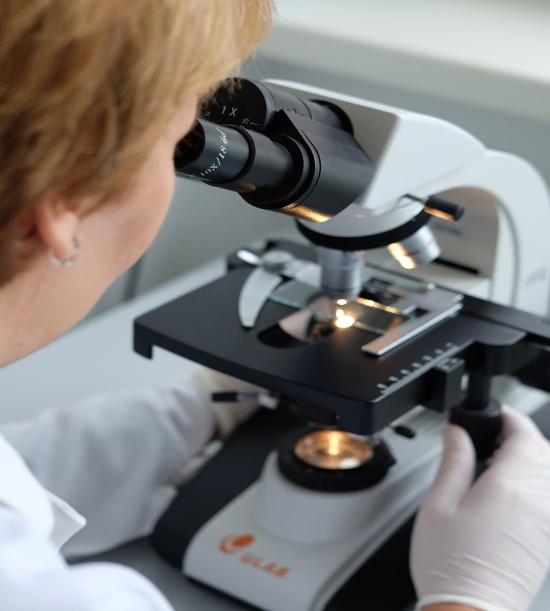A close-up of a researcher looking through a compound microscope, adjusting lenses with gloved hands in a lab setting.