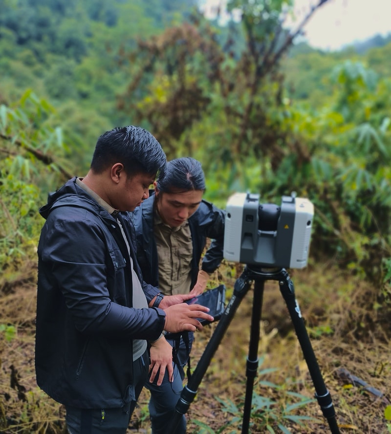 Two people stand in a lush outdoor area with a surveying device on a tripod, one operating a handheld tablet while the other leans in, focused on the screen.