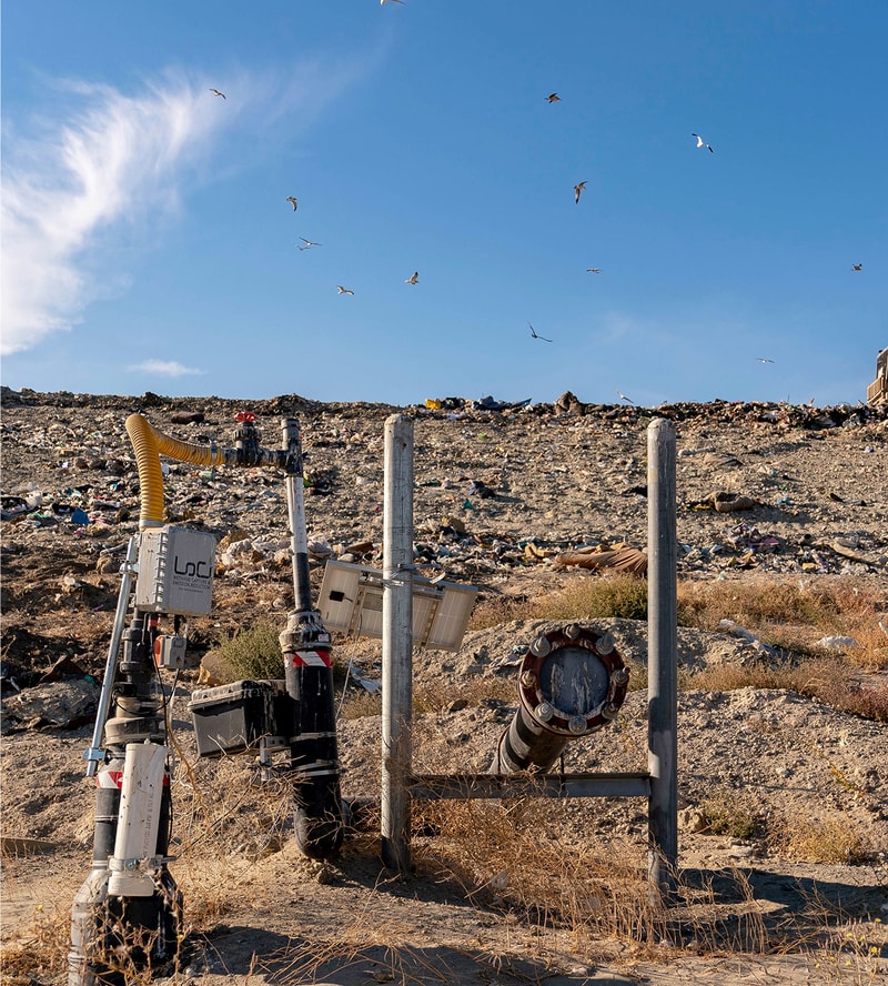Industrial equipment and pipes in a barren, trash-strewn landscape under a blue sky with seagulls flying overhead.