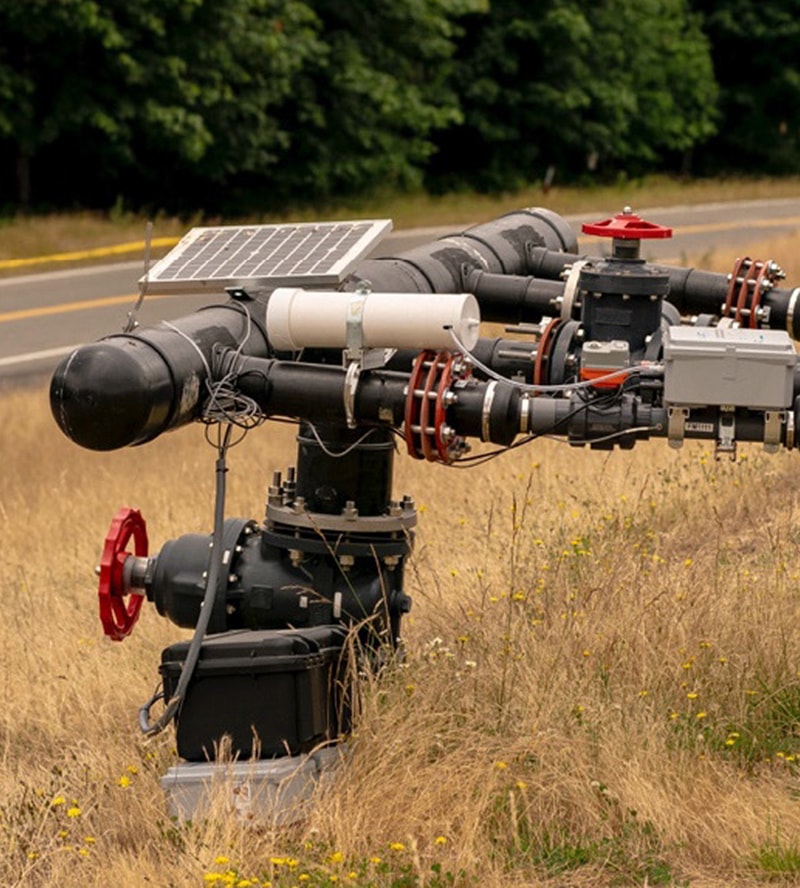 A gas pipeline valve assembly with black pipes, red wheel handles, a solar panel, and electrical components in a grassy field by a road.