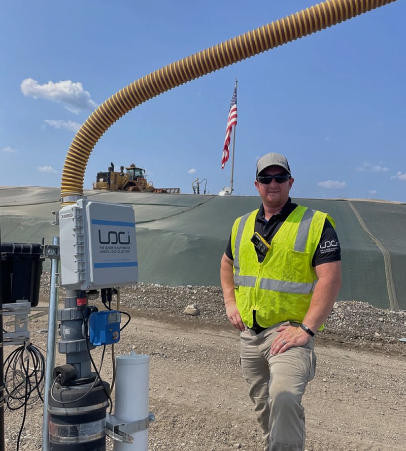 Worker in high-visibility vest and cap standing next to industrial machinery outdoors, American flag in background, blue sky, gravel ground, and coiled yellow conduit above.