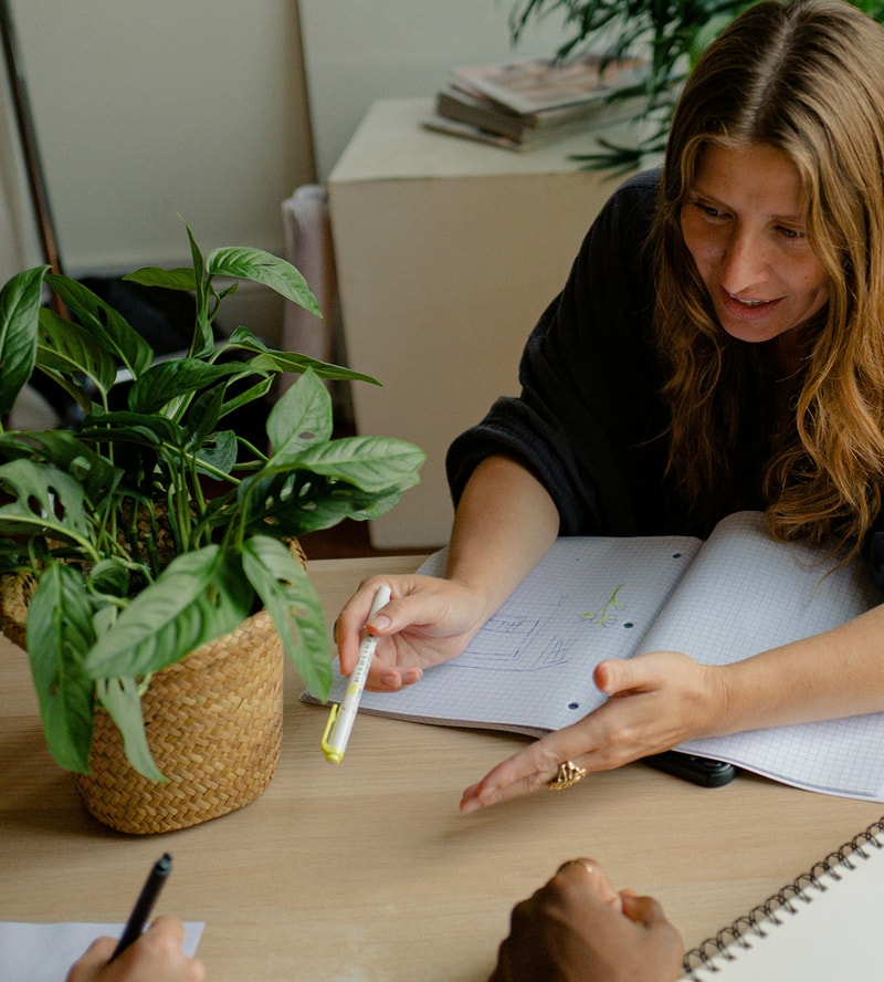 A woman with long hair leans over a notebook, holding a yellow-edged highlighter while discussing plans at a wooden table; a green potted plant sits nearby.