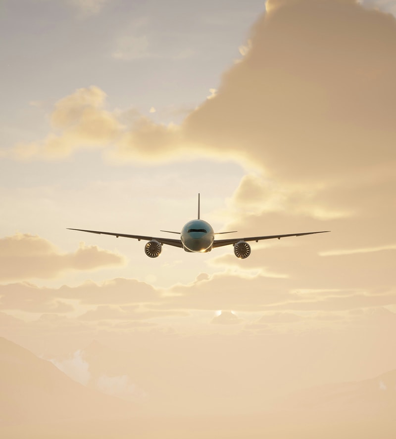 A passenger jet flies toward the camera at sunset, wings wide, engines visible, against a warm, golden sky with soft clouds and distant mountain silhouettes.