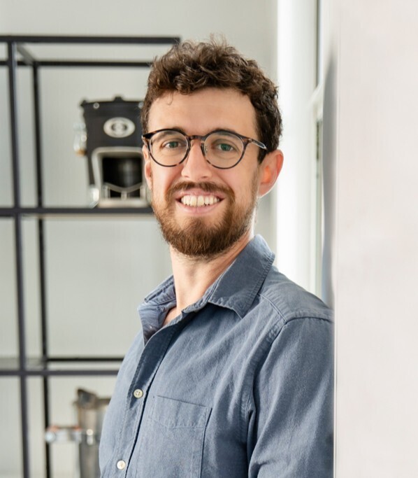 Smiling man with short curly hair, beard, and round glasses stands in a modern office near a metal shelving unit with a coffee machine in the background.