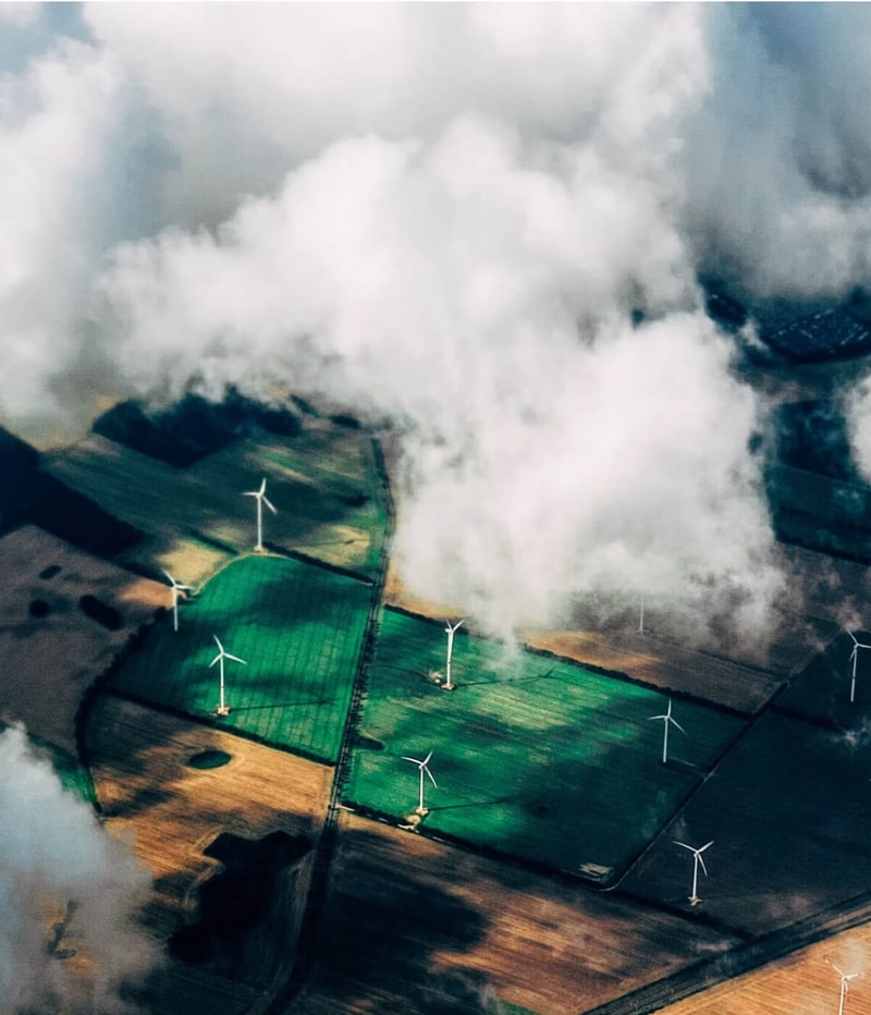 Aerial view of green and brown farmland with several white wind turbines, partially obscured by large white clouds and mist.