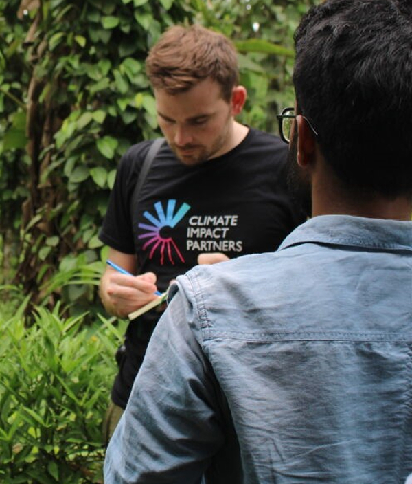 Two men outdoors; one facing slightly away wearing a black T-shirt with "Climate Impact Partners" logo, writing in a notebook while standing among green plants.
