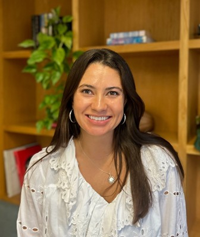Smiling woman with long dark hair wearing a white embroidered blouse and hoop earrings, seated in front of wooden shelves with plants and books.