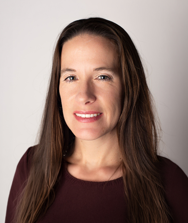 Portrait of a woman with long brown hair, wearing a burgundy top, softly smiling against a light neutral background.