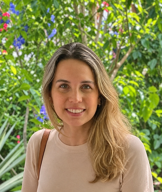A smiling woman with long light brown hair stands outdoors in front of a lush garden with green leaves and colorful flowers.